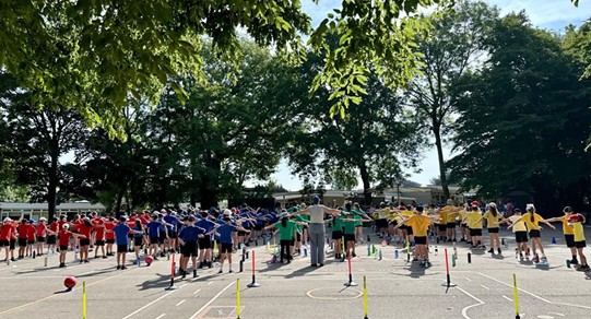 A large group of children, divided into different colored teams (red, blue, green, yellow), are assembled outdoors on a playground or court. They appear to be doing some warm-up exercises or receiving instructions, with an adult in the center leading them. There are cones and markers on the ground, suggesting various activity stations.