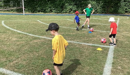 Several young children are practicing or playing with footballs on a grassy area marked with white lines (likely a mini-pitch). They are spread out and appear to be working on individual ball skills or a small-group game.