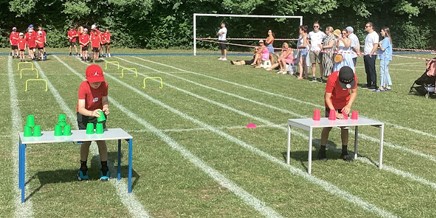 Two children are competing in a cup stacking race on tables set up on a grassy track. In the background, other children in red shirts are lined up, possibly waiting for their turn or watching another event. Spectators are visible behind a barrier.