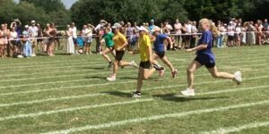 Girls are participating in a running race on a grassy track marked with white lines. Spectators are cheering them on from behind a rope barrier.