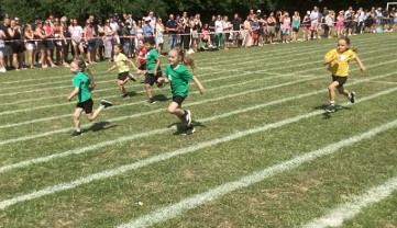 Younger children are running a race on the grassy track. They are spread across multiple lanes, and adult spectators are watching from the side.