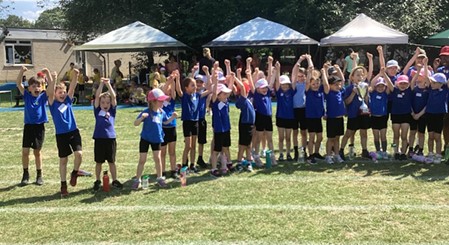 A group of children in blue shirts are gathered together, many with their arms raised in celebration or excitement. They look like another team, possibly posing after an event or getting ready for one. A trophy is visible among them. Tents are in the background.
