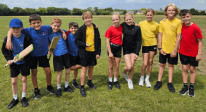 10 Year 6 pupils in PE kit line up for a photo on a sports field. Two are holding cricket bats and one a ball. Three are dressed yellow tops, two in red, one in black and four in blue.