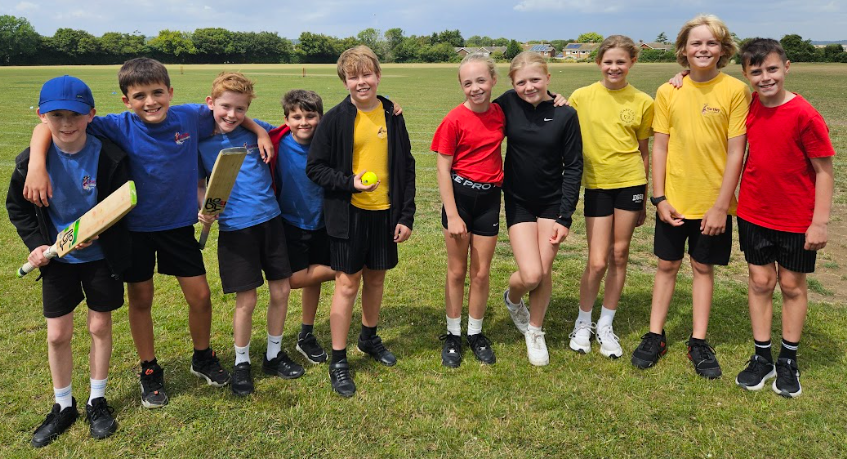 10 Year 6 pupils in PE kit line up for a photo on a sports field. Two are holding cricket bats and one a ball. Three are dressed yellow tops, two in red, one in black and four in blue.