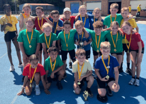 20 pupils in PE kit in varying colours pose for a photo in three rows on a school playground. All of them have medals and there are two trophies on show.