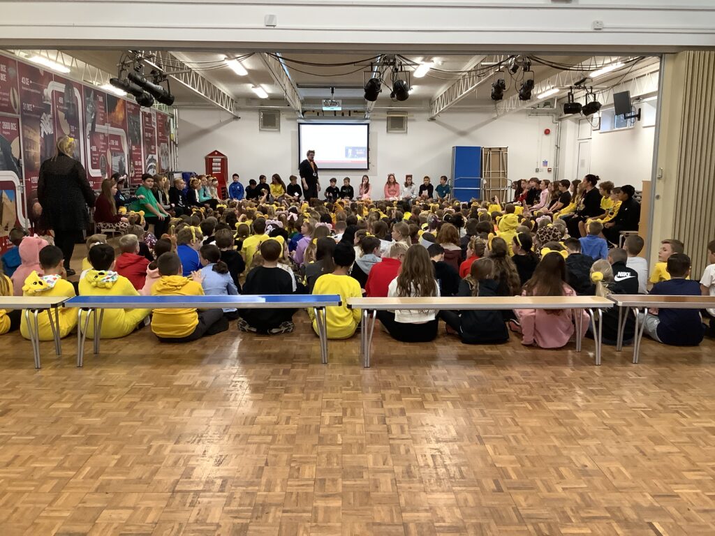 A wide shot of a school assembly hall with a wooden parquet floor, where a large group of children, many wearing yellow tops or bear-ear headbands, are sitting on the floor and benches, facing a stage. On the stage, several adults are sitting and standing, addressing the children.