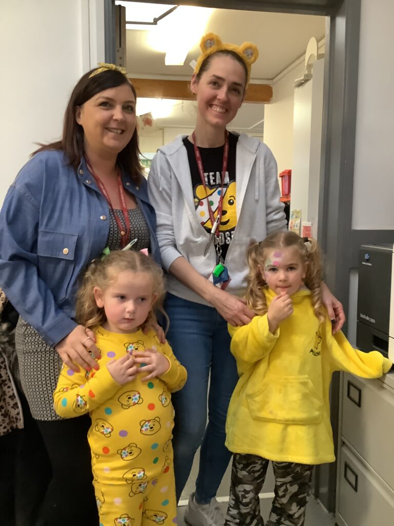 Two adults and two young children smile and pose for a photo in an indoor setting, possibly an office. The children are in the front, and the adults stand behind them. Both children and one adult wear yellow-themed clothing or bear-ear headbands. A filing cabinet is visible to the right.