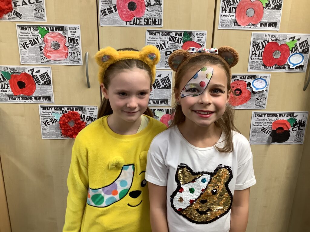 Two children stand side-by-side smiling for a photo. Both wear bear-ear headbands. The child on the left wears a yellow fleece. The child on the right wears a white t-shirt with a sequined bear and has colorful spots painted over one eye. The wall behind them is covered with student art projects about 'VE-DAY' and poppies.