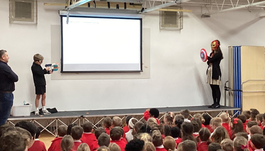 On a school stage, a child holds a toy blaster and a presenter wears an Iron Man mask and holds a Captain America shield during a presentation. A large audience of students in red uniforms watches from below.