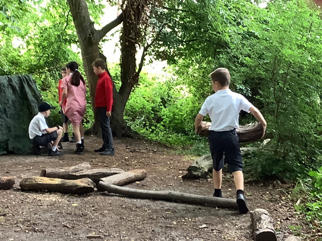 The image shows a group of children in a wooded outdoor area. On the left side of the frame, four children are gathered near what looks like a green tarpaulin or makeshift shelter. One child in a white shirt and black shorts is crouching, while the others are standing. In the foreground, a child in a white shirt and dark shorts is walking away from the viewer, carrying a large piece of wood or a log. There are several other logs scattered on the ground. The background is filled with lush green foliage, indicating a natural, possibly forested, environment.