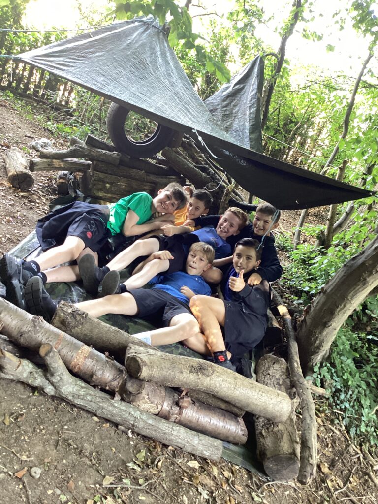 This image captures a group of people, appearing to be engaged in an outdoor activity, likely bushcraft or shelter building, in a wooded environment. In the foreground, there are several individuals gathered closely together, smiling and looking at the camera. They are lying or sitting on what appears to be a tarp or ground cover, surrounded by logs and branches that form part of a structure. Above them, a makeshift shelter has been constructed using tarpaulins draped over branches and possibly an old tire, providing cover. The background shows more trees and foliage, indicating a natural, outdoor setting.