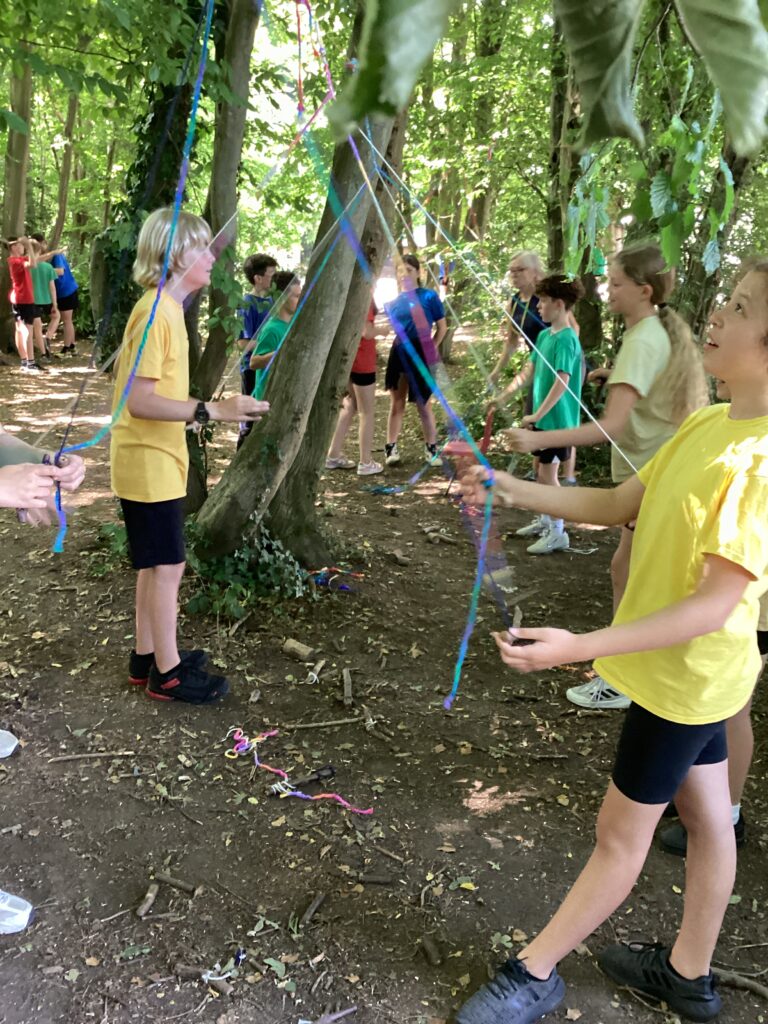 This picture shows a group of children and adults participating in an outdoor activity, possibly a "low ropes course" or a similar team-building exercise. They are in a wooded area, surrounded by trees and foliage, with sunlight dappling through the leaves. Several people are holding onto colorful ribbons or ropes, which appear to be stretched between trees or held by others. The children are dressed in various casual clothing, including t-shirts and shorts. The ground is covered with dirt, leaves, and some scattered twigs. The overall impression is one of an active and engaging outdoor group activity.