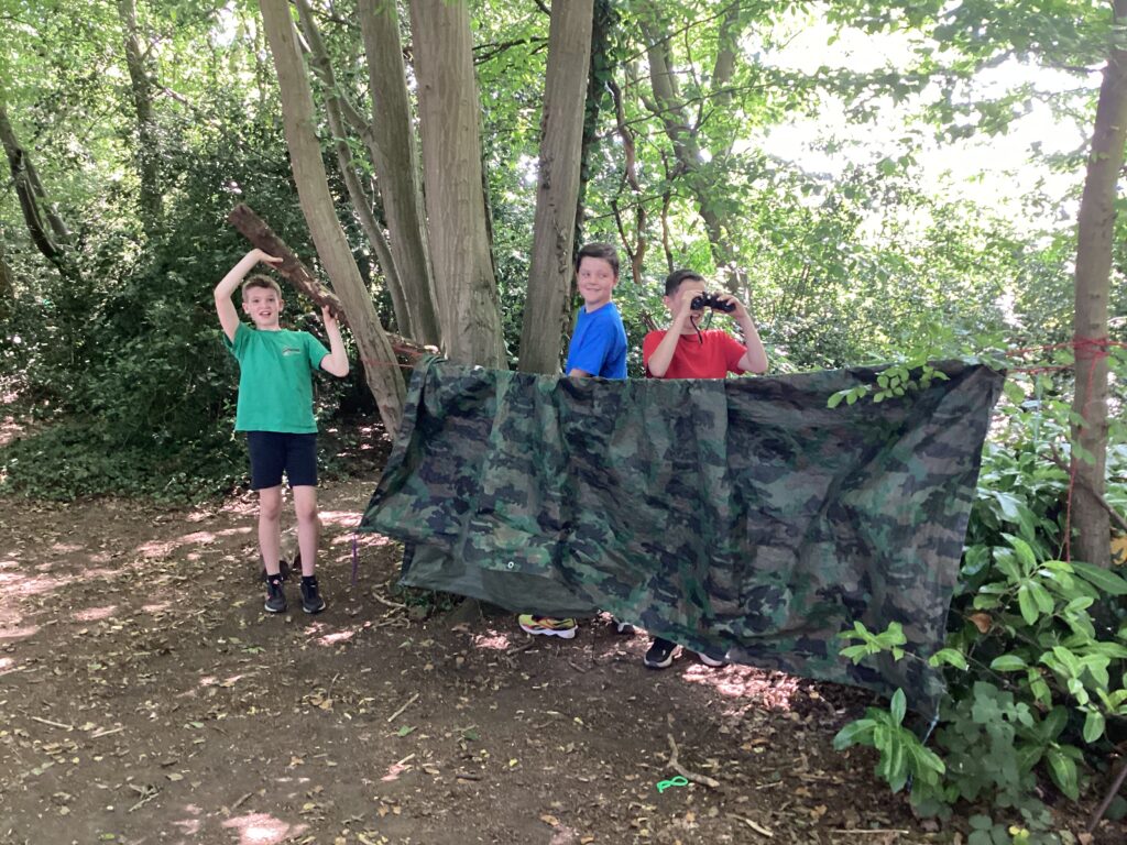 This picture shows three individuals in a wooded outdoor setting. They appear to be engaged in an activity related to building a shelter or fort. In the foreground, a camouflage-patterned tarpaulin or sheet is strung up between trees, forming a partial barrier or hideout. To the left, one person is standing and holding a long stick or branch. In the middle, another person is peeking out from behind the camouflage material. To the right, a third person is also peeking out from behind the material, holding a pair of binoculars to their eyes. The ground is covered with dirt and some foliage, and trees are visible in the background, suggesting a natural environment.