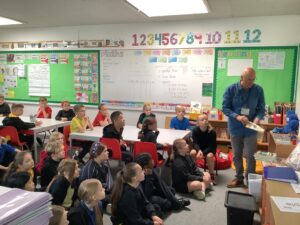 Setting: It's a brightly decorated classroom. The walls feature educational posters, including a large number line, a "Maths" board with place value and inequality symbols (> and < signs), and a "Reading" area. The walls have a prominent green border. Students: There are numerous students, likely in the junior years (Key Stage 2), seated in two main groups: A group of children are sitting on the floor near the front, facing the teacher. Another group is seated at white tables in the middle and back of the room. Most students are wearing black or dark uniform tops (sweatshirts or polo shirts). Teacher: An older man, presumably the teacher or a visiting adult, is standing on the right side of the frame. He is holding a book and appears to be reading to the class or introducing a story/topic. Activity: The arrangement—students gathered on the floor and the teacher holding a book—suggests a story time, a reading comprehension session, or the introduction of a new topic, possibly in literacy or a themed subject. Despite the prominent "Maths" board, the immediate activity being led by the teacher seems to be centered around reading. Overall, it's a typical scene of children attentively participating in a lesson being delivered by their teacher.