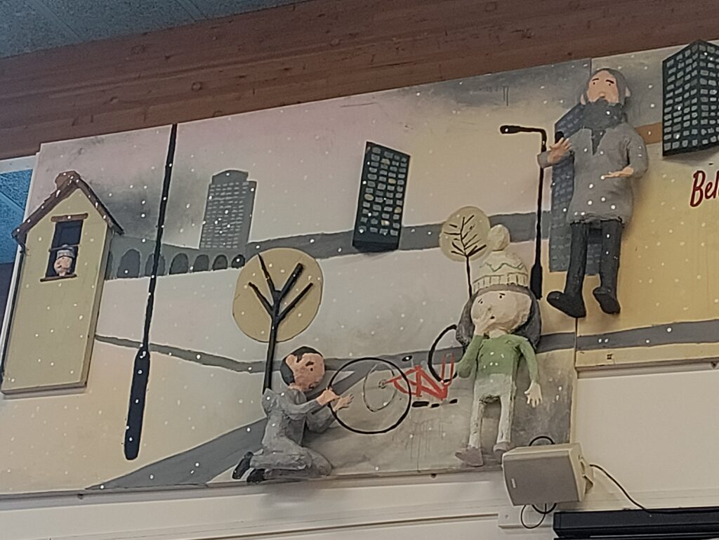 This section of a mural has a cool, monochromatic palette dominated by black, white, and grey, suggesting a winter or cold urban setting with falling snow. Figures include a small figure looking out of a window in a small, peaked-roof house, a man kneeling to tend to a bicycle wheel, and another figure in a knit hat standing nearby. A taller adult figure wrapped in a scarf stands further right. All raised figures are sculpted and wear thick clothing, reinforcing the cold weather theme.