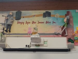 This is the heart of the mural, bridging the cold and warm sections and containing the main text. The central scene depicts two small figures standing near each other with potted flowers between them, suggesting an act of connection or sharing.