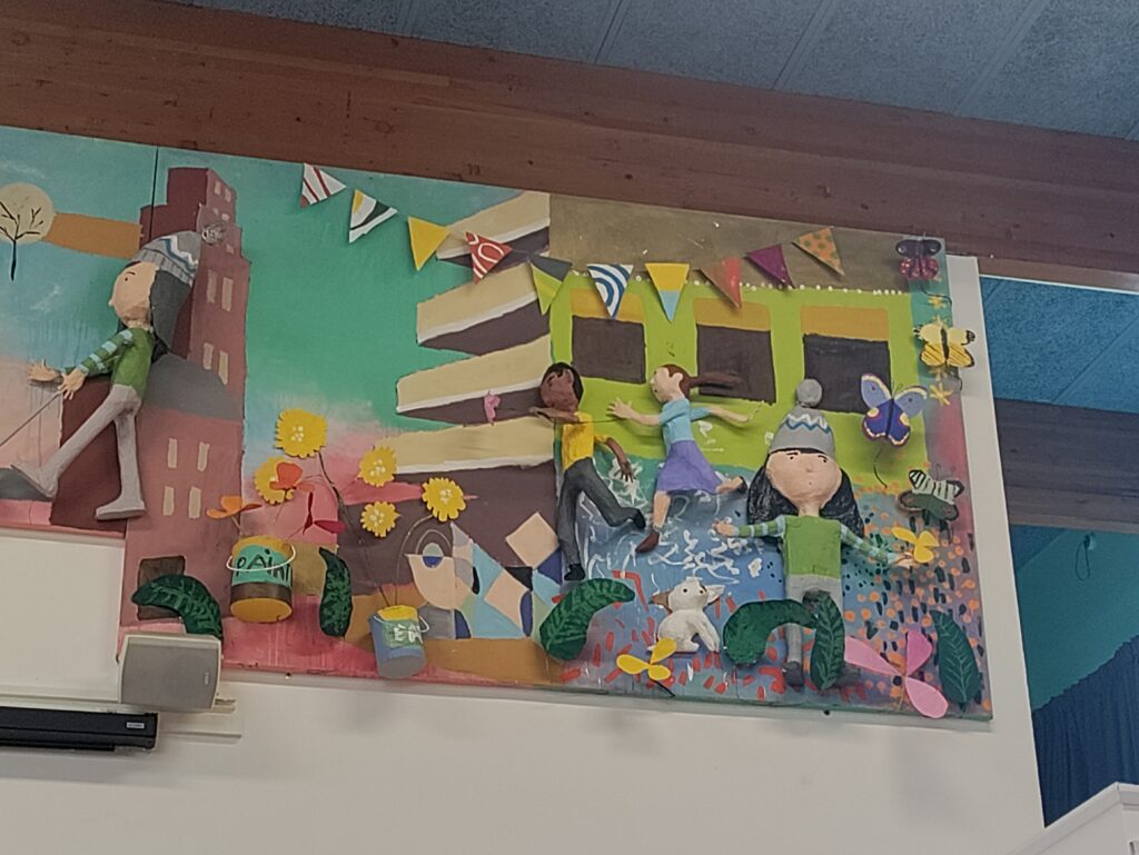 This part of the mural is characterized by a warm, vibrant palette featuring teal, red, yellow, and green, suggesting a lively, summer or festive atmosphere. Figures are sculpted and appear to be dancing or running with their arms outstretched, reflecting joy and movement. They are diverse in appearance and clothing.