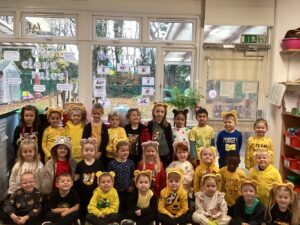 A group of young children pose for a photo in three rows inside a classroom. Many are wearing yellow clothing and bear-ear headbands. Behind them is a large window with classroom displays, including a 'Welcome' sign, and trees are visible outside.