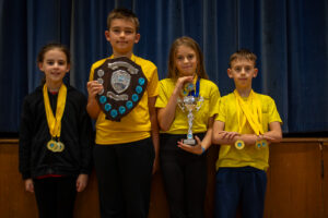 four young children wearing yellow tops posing in front of a blue backdrop holding trophies and medals