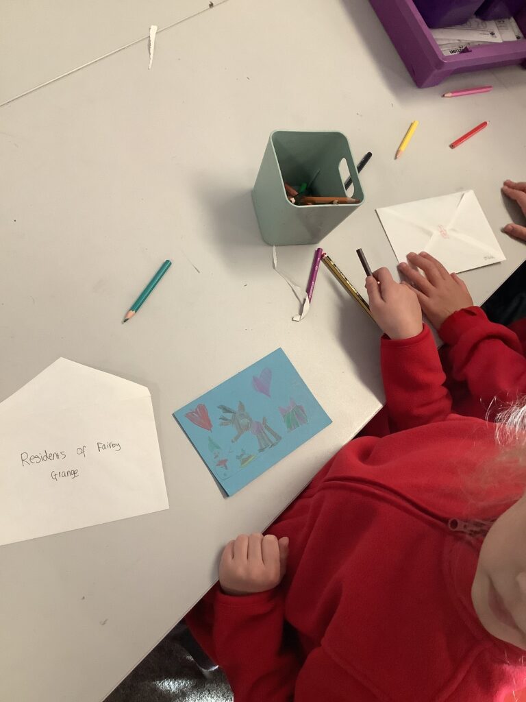 A high-angle shot of a child in a red sweatshirt coloring a blue Christmas card featuring a drawing of a reindeer. An envelope nearby is addressed to "Residents of Fairby Grange," and several colored pencils are scattered across the white table.