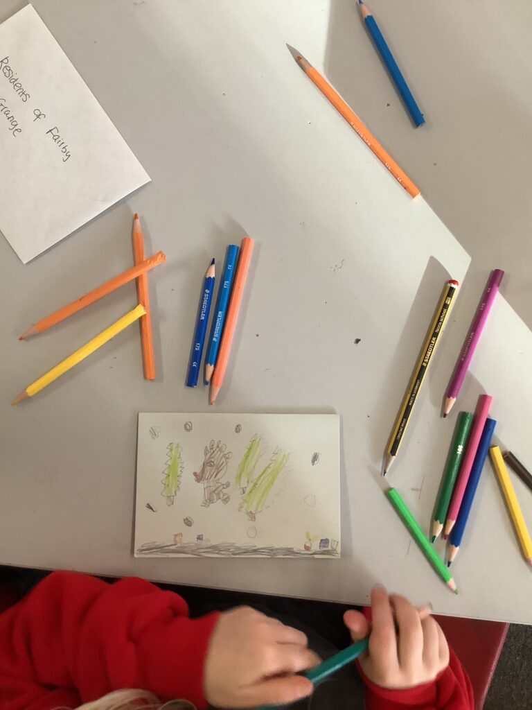 A top-down view of a child's hands holding a green colored pencil over a white card with a drawing of a reindeer and Christmas trees. The table is covered with various colored pencils and an envelope addressed to "Residents of Fairby Grange."