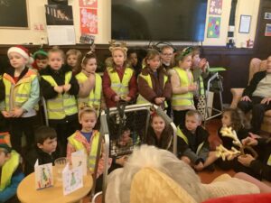A group of school children in high-visibility vests and holiday headbands standing together in a common room. Several children are seated on the floor in front of them, and two handmade Christmas cards are propped up on a small table.