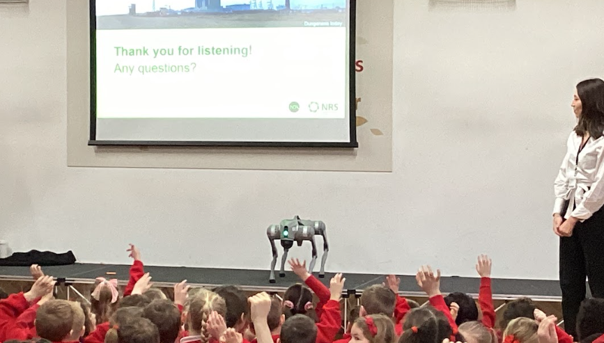 A presenter stands on a stage next to a four-legged robotic dog, while a screen behind them displays "Thank you for listening! Any questions?" and the NRS logo. Many children in the foreground have their hands raised.
