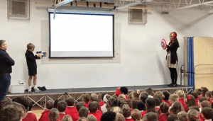 On a school stage, a child holds a toy blaster and a presenter wears an Iron Man mask and holds a Captain America shield during a presentation. A large audience of students in red uniforms watches from below.
