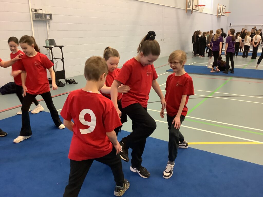 Four children in red t-shirts practice a synchronized movement on a blue gym mat. One child in the center balances on one leg while others provide support or mimic the stance.
