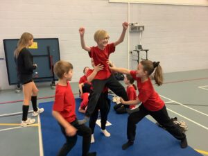 A group of seven children in red shirts pose for a celebratory photo. They are holding one teammate in a horizontal split position while others cheer and smile toward the camera.