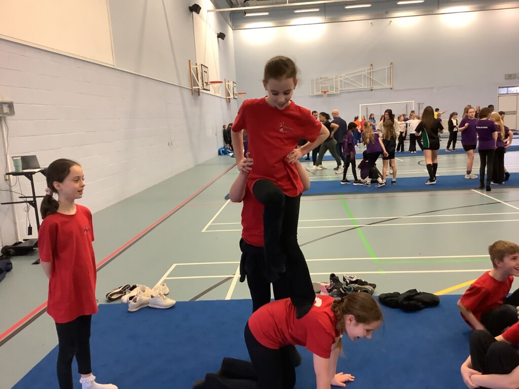 Two children in red shirts practice a vertical balance move. One child is on their hands and knees acting as a base, while another child stands carefully on their back, supported by a third teammate.