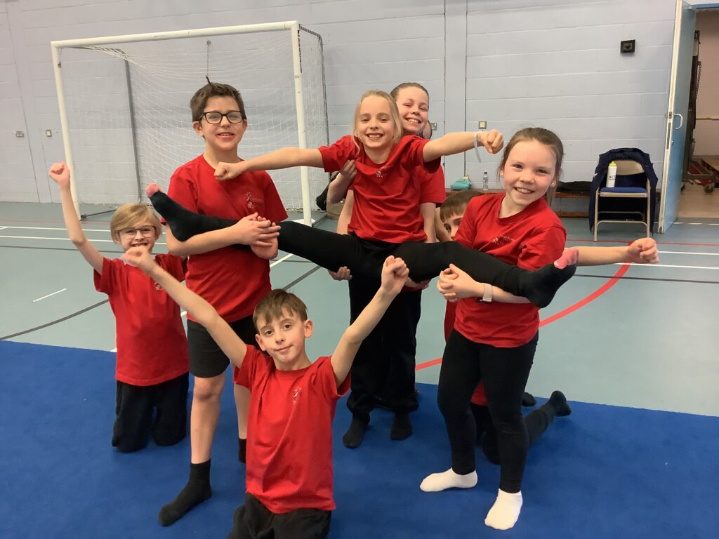 A group of seven children in red shirts pose for a celebratory photo. They are holding one teammate in a horizontal split position while others cheer and smile toward the camera.