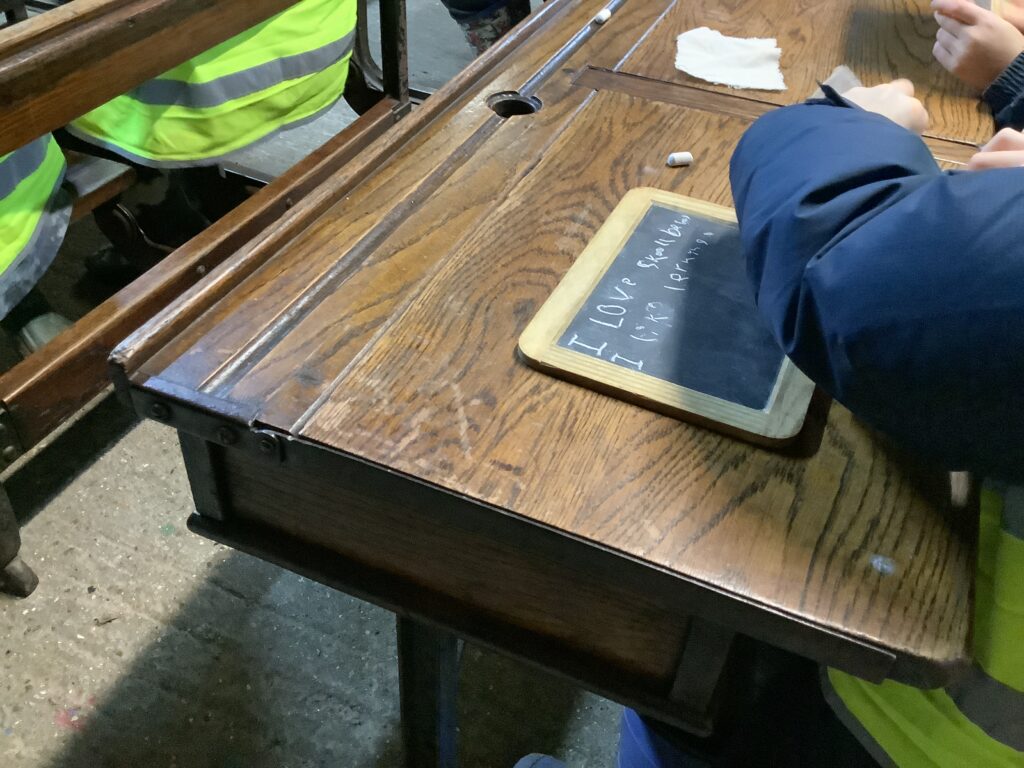 A child sits at an old-fashioned school desk using chalk to write on a small slate board.
