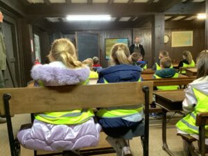 Children in high-vis vests sit on wooden benches listening to a teacher in a dark, traditional classroom.