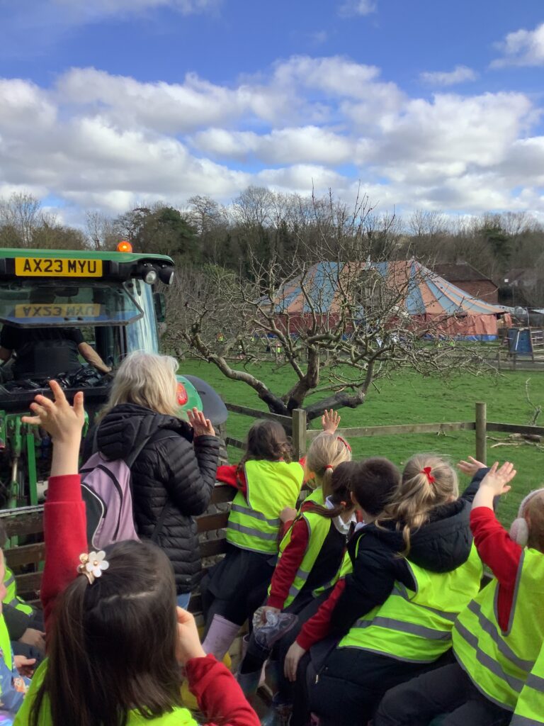 A group of children in high-vis vests sit on a trailer being pulled by a tractor, waving.