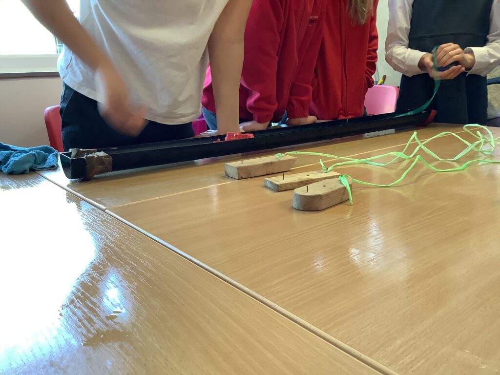 Several students stand at a table, leaning over a long, black track and wooden blocks with green strings attached.