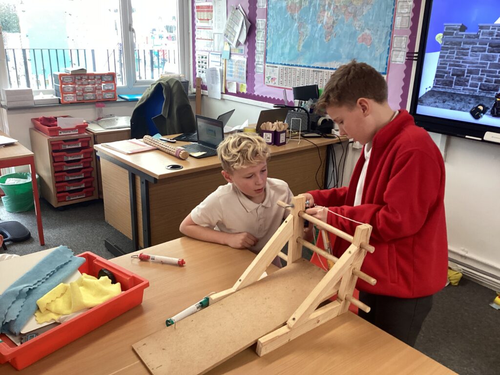 Two students collaborate in a classroom setting to adjust a wooden frame and pulley-like mechanism on a wooden board.
