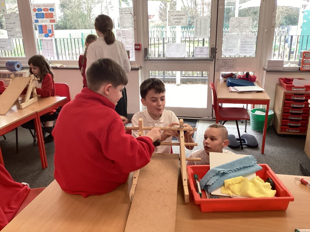Three students work together at a desk to assemble a wooden structure that resembles a small ramp or bridge.