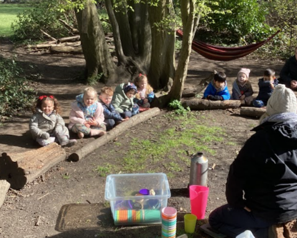 Children sitting in a circle, with logs in a wooded area. A teach is in the middle with a flask and cups.