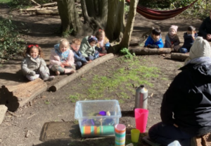 Children sitting in a circle, with logs in a wooded area. A teach is in the middle with a flask and cups.
