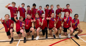A group of 17 children posing in a sport hall holding gold medal and holding up one finger.