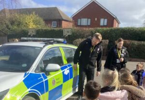 Two police officers stand in front of a police car on a school playground talking to a group of young pupils