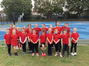 A group of primary school girls in matching red T-shirts pose in two rows on grass in front of a fenced multi-use games area with a blue court surface. Most wear black leggings or shorts with trainers, and several smile with their hands clasped. One girl in the front row rests her foot on a red and blue football, and houses and trees are visible behind the court.
