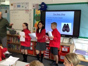 Three pupils in red jumpers read from sheets of paper in front of a classroom screen showing a picture of a pair of shoes.