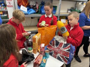 Primary pupils in red school jumpers sort through donated items on a table, with one pupil lifting a yellow item out of a large orange bag. Food packets and boxes are spread across the table in front of them.
