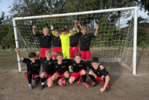 A primary boys' football team in black and red kit celebrate in front of a goal, with their goalkeeper in yellow at the centre.