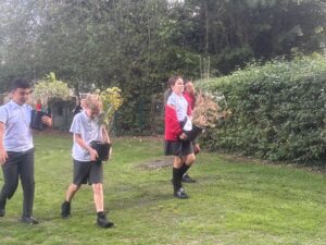 Four pupils in school uniform carry potted shrubs across a grassy area of the academy grounds, with hedges and trees behind them.